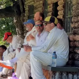 Cricket players in white watching a match from a wooden pavilion steps